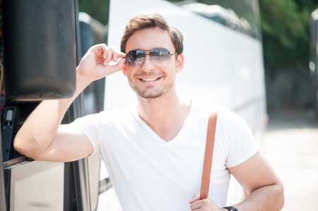 Cheerful man is standing near a bus and waiting for departure. He is wearing sunglasses and smiling. The man is holding a rucksackの写真素材