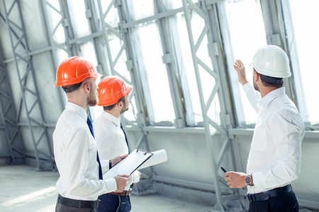 Attractive architect is explaining to the workers the plan of construction with seriousness. He is pointing his arm sideways. His colleagues are looking forward with concentrationの写真素材