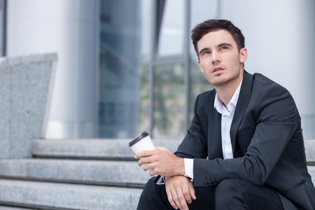 Cheerful businessman is sitting on steps outdoors. He is drinking coffee and relaxing. The man is looking up dreamingly. Copy space in left sideの写真素材