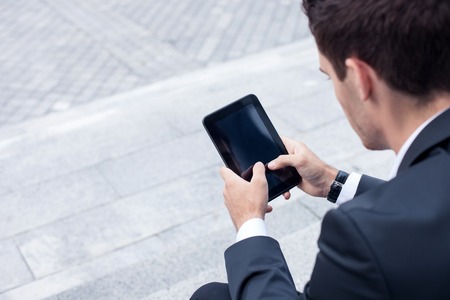 Successful male worker is holding a tablet in his hands. He is touching it and looking at it with joy. The man is sitting on steps. Copy space in left sideの写真素材