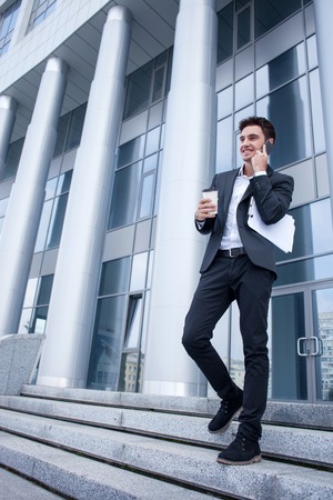 Successful man in suit is moving on steps outdoors. He is talking on the phone and smiling. The man is holding documents and a cup of coffee. Copy space in left sideの写真素材