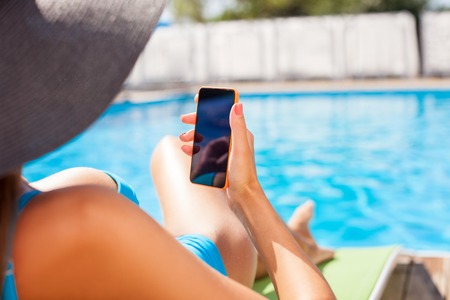 Close up of body of beautiful woman is lying near a swimming pool. She is sunbathing. The woman is holding a mobile phone and messaging to someoneの写真素材