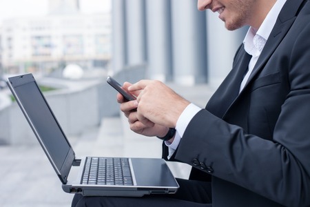 Close up of cute businessman is sitting on steps and holding a laptop on his knees. The man is using a mobile phone for communication. He is touching it with joy and smilingの写真素材