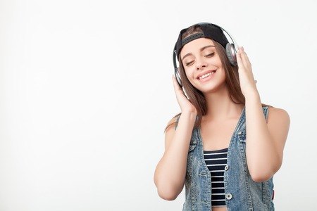 Cheerful hipster girl is listening to music from headphones. She is touching it with pleasure and smiling. Her eyes are closed with enjoyment. Isolated on background and copy space in left sideの写真素材