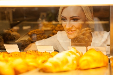 Beautiful blond girl is looking at the display of appetite baked products with temptation. She is standing in bakery and smilingの写真素材