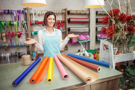 Beautiful saleswoman is standing at the counter in her flower shop. She is raising her arms sideways and presenting a wide selection of wrapping paper. The woman is smilingの写真素材