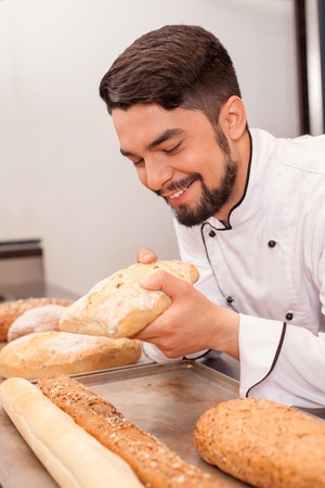Cheerful male baker is holding pastry and checking its quality. He is standing in bakery and...