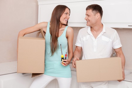 Pretty husband and wife are packing their staff for moving in another house. They are carrying cardboard boxes and smiling. The woman is holding screwdriver and pliers. She is looking at man with joyの写真素材