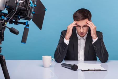 Professional reporter has a terrible headache. He is sitting at the table and touching his temples. The man is looking down at the documents with frustration. Isolated on blue backgroundの写真素材