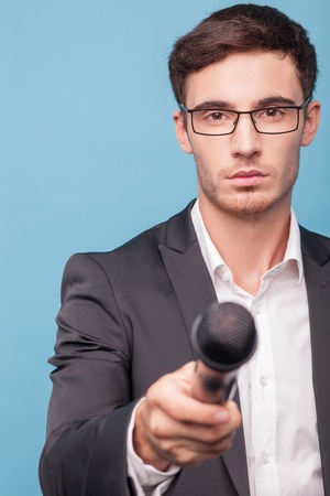 Cheerful reporter is interviewing someone. He is holding a microphone and proposing someone to tell something. The man is looking forward seriously. Isolated on blue backgroundの写真素材