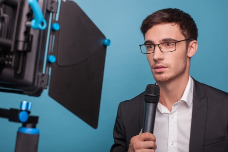 Attractive reporter is holding a microphone and telling news. The man is looking at the camera seriously. He is wearing suit and eyeglasses. Isolated on blue backgroundの写真素材