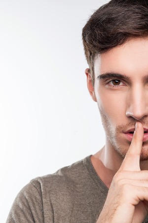 Close up of handsome guy is asking for silence. He is raising his index finger to his lips secretly. The guy is looking at the camera with seriousness. Isolated on backgroundの写真素材