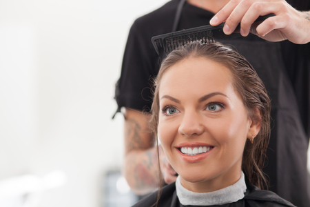 Beautiful woman is sitting and smiling in beauty shop. The hairdresser is combing her wet hair with concentration. Copy space in left sideの写真素材