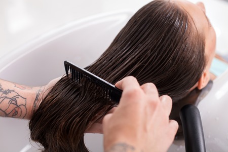 Close up of arms of hairstylist combing female hair with balsam. The woman is leaning her head on the sink and relaxing. Her eyes are closed with enjoymentの写真素材