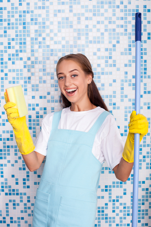 Cheerful female cleaner is doing clean-up in a bathroom. She is holding a mop and a sponge in her hands. The woman is looking at the camera and smilingの写真素材