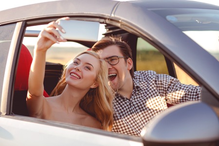 Cheerful young man and woman are sitting in car and making selfie. They are looking at the mobile phone and laughing. The man is driving a vehicleの写真素材