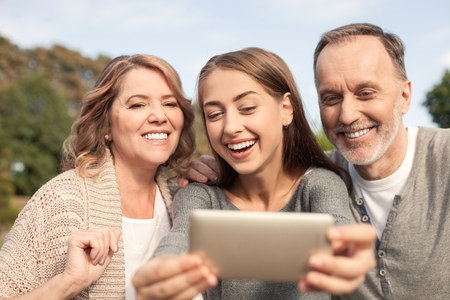 Pretty mature family is making selfie with joy. They are sitting in park and embracing. The family is looking at the mobile phone and smiling. The young woman is holding telephoneの写真素材
