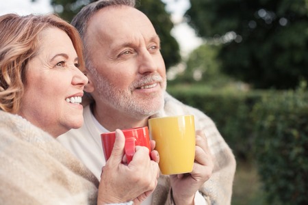 Cheerful mature husband and wife are drinking tea outdoors. They are sitting and embracing. The man and woman are warming with quilt. They are smilingの写真素材