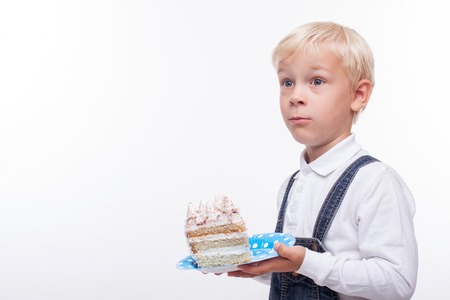 Cute boy is holding a plate with piece of cake. He is standing and looking aside with surprise. Isolated and copy space in left sideの写真素材