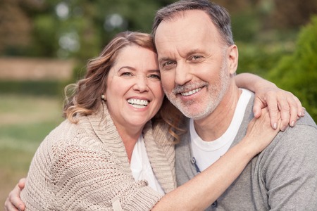 Pretty old husband and wife are sitting in park. They are embracing and smiling. The man and woman are looking at the camera with happinessの写真素材