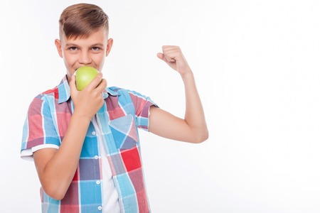 Cheerful male teenager is eating an apple with joy. He is flexing his bicep and showing his strength. The schoolboy is looking at the camera happily. Isolated and copy space in right sideの写真素材