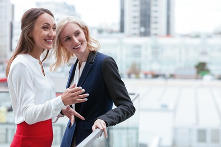 Cheerful businesswomen are standing near the border of building and talking. They are looking forward and smiling. The brunette woman is gesturing with inspiration. Copy space in right sideの写真素材