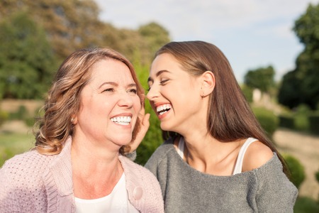 Attractive young woman is talking with her mother happily. They are standing in park and embracing. The mother and daughter are laughingの写真素材