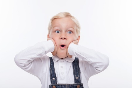 Cute small boy is looking forward with shock. He is standing and touching his face with amazement. His mouth is wide open. Isolated on backgroundの写真素材