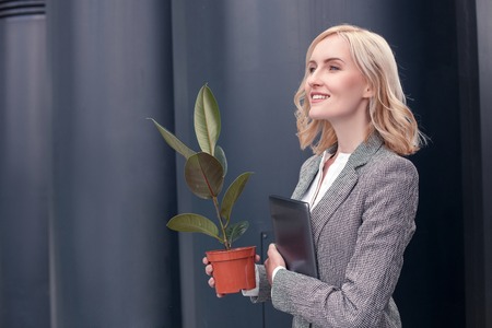 Cheerful businesswoman is standing and smiling. She is holding a plant and a laptop. The blond lady is looking forward proudly. Copy space in left sideの写真素材