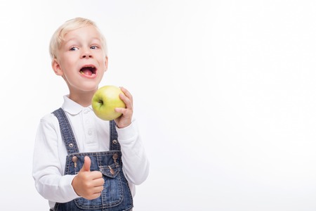 Pretty hungry boy is eating a healthy apple with happiness. He is standing and giving thumb up with satisfaction. The boy is smiling. Isolated and copy space in right sideの写真素材