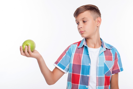 Beautiful boy is holding an apple and looking at it with appetite. He is standing in colored shirt. Isolated on backgroundの写真素材