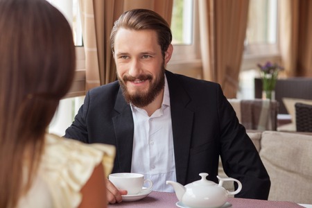 Beautiful woman and man are dating in restaurant. They are sitting opposite each other and smiling. The bearded man in suit is drinking teaの写真素材
