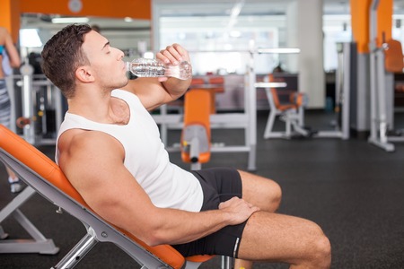 Handsome fit athlete is drinking water with enjoyment. He is sitting on bench and resting in gym. The man closed his eyes with relaxationの写真素材