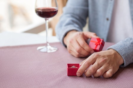 Close up of male hands holding a small box of gold ring. He is gifting it to his girlfriend. The man is sitting at the table and drinking red wineの写真素材