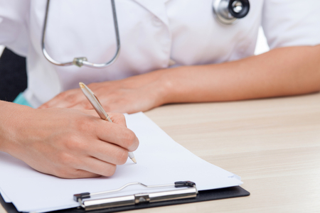 Close up of hands of nurse writing some data into the folder of papers. The woman is sitting at the table. She is wearing a stethoscopeの写真素材