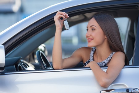 Attractive businesswoman is sitting at steering wheel of her car. She is holding a key and looking at it with happiness. The lady is driving and smilingの写真素材