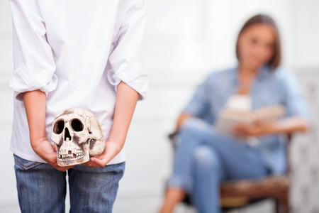 Close up of body of boy holding a toy skull. He is hiding it behind his back from his parent. The woman is sitting in chair and reading book. Focus on back of sonの写真素材