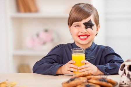 Pretty girl is drinking orange juice in the kitchen. She is ready for celebration Nut-Crack Night. The child is looking forward and smiling. There are spooky cookies and skull on the tableの写真素材