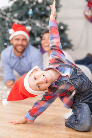 Cheerful small boy is sitting on flooring and posing near New Year tree. He is wearing red hat. His parents are lying behind him and looking at son with joy. They are laughingの写真素材