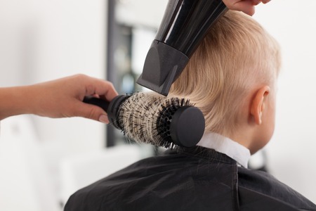 Close up of hands of hairdresser. The woman is standing near a blond boy. She is holding a hairdryer and the comb. She is making a hairstyle to childの写真素材