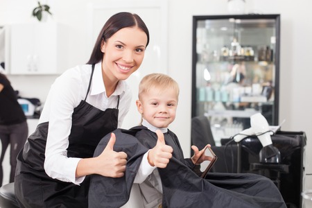 Cute hairdresser is making a haircut for child. The boy is sitting and holding a tablet. The woman and child are giving thumbs up. They are smiling and looking forward happilyの写真素材