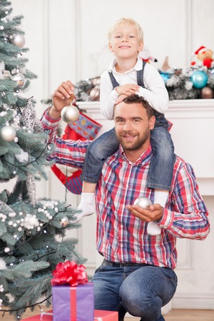 Cheerful father and son are decorating Christmas tree. They are looking at the camera and smiling. The parent is holding boy on his shoulders and spheres in hands. The boy is sitting with joyの写真素材