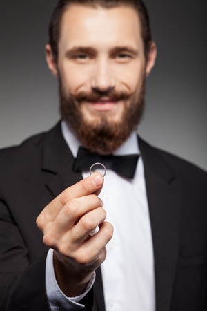 Cheerful businessman with beard is wearing suit. He is standing and holding a ring. The man is making proposal and smiling. Isolated on black backgroundの写真素材