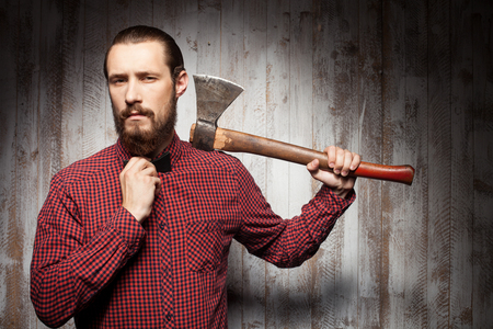 Attractive lumberjack with beard is holding a hatchet. He is standing and adjusting the bow-tie on his neck. The man is looking at the camera self-confidently. Copy space in right sideの写真素材