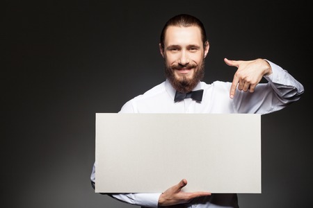 Cheerful bearded man is showing white empty billboard. He is pointing his finger at it and smiling. The man is standing and looking at the camera happily. Isolated and copy space in left sideの写真素材