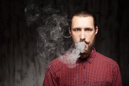 Handsome bearded man is standing and breathing out smoke. He is looking forward seriously. Copy...
