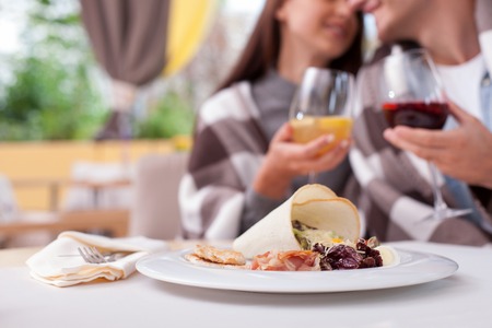 Close up of cute man and woman sitting at the table in cafe. They are drinking cocktail and smiling. They are embracing with quilt on their shoulders. Focus on a plate of saladの写真素材