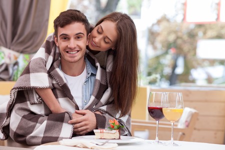 Beautiful man and woman are resting in restaurant. The man is sitting and looking at the camera happily. His girlfriend is standing and embracing him. They are smiling and warming with quiltの写真素材