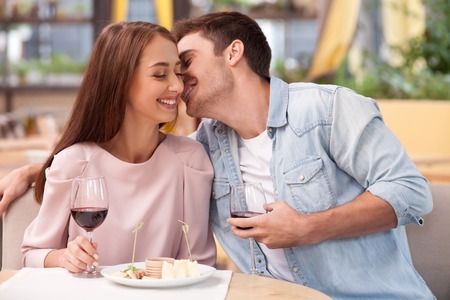 Beautiful young loving couple is dating in restaurant. They are sitting near each other. The guy is embracing his girlfriend. They are drinking wine and smilingの写真素材