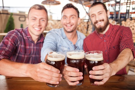 Cheerful men are drinking beer in pub. They are sitting at counter and smiling. The guys are clinking glasses and looking at camera happily. Focus on three glasses of beerの写真素材